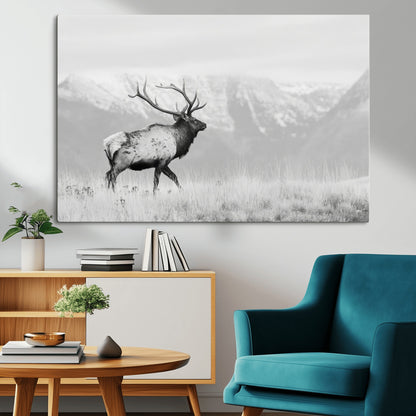 Black and white photograph of a majestic elk standing in a grassy field with snow-capped mountains in the background, creating a bold and rustic wilderness scene.