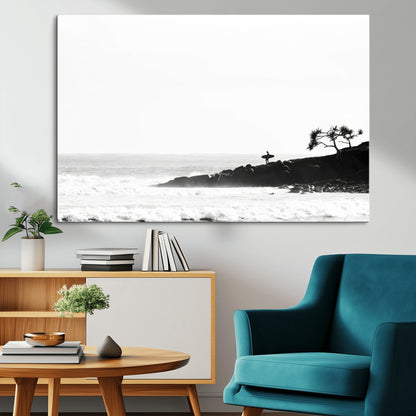Black and white photo of a surfer standing on rocky cliffs, gazing at the waves, embodying the spirit of adventure and solitude.