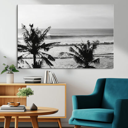 Black and white view of ocean waves crashing on a tropical coastline, framed by silhouetted palm trees under a vast, open sky.