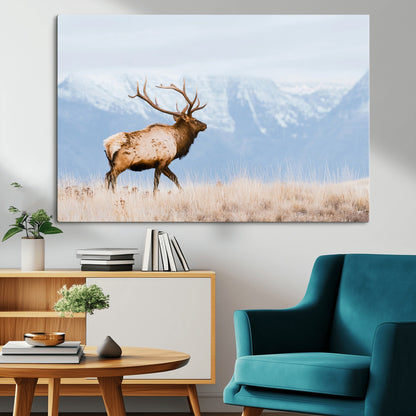 An elk walking on snowy grassland with majestic mountains in the background, ideal for rustic lodge or cabin wall decor.