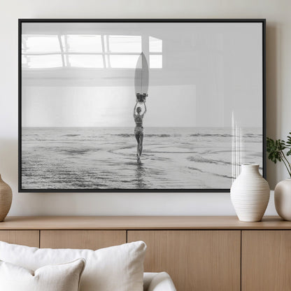 Black and white image of a female surfer holding a surfboard above her head while walking toward the ocean, creating a minimalist and bold coastal aesthetic.