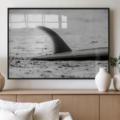A black and white close-up of a surfboard fin resting on the sandy shore, highlighting minimalist beach aesthetics and classic surf culture.