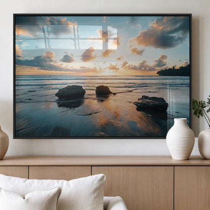 A serene beach at sunset with vibrant clouds, calm water, and rocks in the foreground reflecting the warm colors of the setting sun.