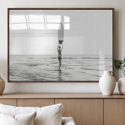 Black and white image of a female surfer holding a surfboard above her head while walking toward the ocean, creating a minimalist and bold coastal aesthetic.