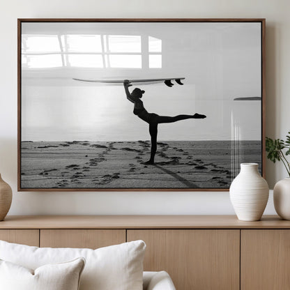 A woman balances a surfboard on her head while striking a yoga pose on the beach, silhouetted against a calm ocean horizon.