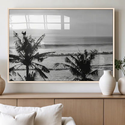 Black and white view of ocean waves crashing on a tropical coastline, framed by silhouetted palm trees under a vast, open sky.