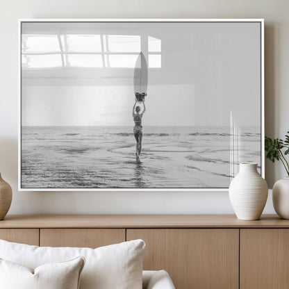Black and white image of a female surfer holding a surfboard above her head while walking toward the ocean, creating a minimalist and bold coastal aesthetic.