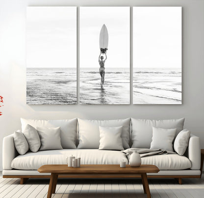 Black and white image of a female surfer holding a surfboard above her head while walking toward the ocean, creating a minimalist and bold coastal aesthetic.