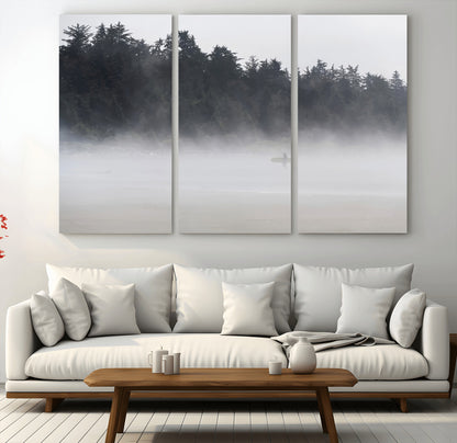 A lone surfer carrying a board through a misty shoreline, surrounded by dense trees and fog, creating a peaceful and mysterious coastal scene.