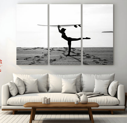 A woman balances a surfboard on her head while striking a yoga pose on the beach, silhouetted against a calm ocean horizon.