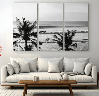 Black and white view of ocean waves crashing on a tropical coastline, framed by silhouetted palm trees under a vast, open sky.