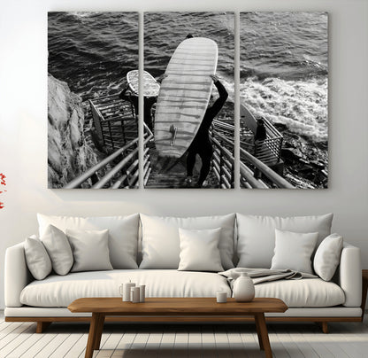 Black and white photo of surfers carrying boards down a rugged cliffside stairway, capturing a bold and adventurous coastal scene.