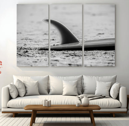 A black and white close-up of a surfboard fin resting on the sandy shore, highlighting minimalist beach aesthetics and classic surf culture.