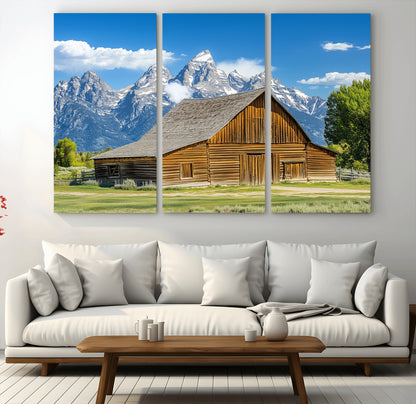 A rustic wooden barn sits in front of towering mountain peaks under a clear blue sky, perfect for farmhouse or cabin style interiors.