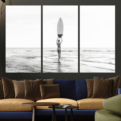 Black and white image of a female surfer holding a surfboard above her head while walking toward the ocean, creating a minimalist and bold coastal aesthetic.