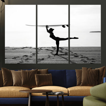 A woman balances a surfboard on her head while striking a yoga pose on the beach, silhouetted against a calm ocean horizon.