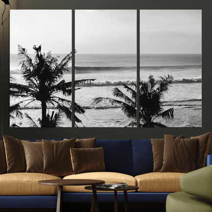 Black and white view of ocean waves crashing on a tropical coastline, framed by silhouetted palm trees under a vast, open sky.