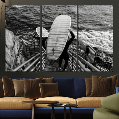 Black and white photo of surfers carrying boards down a rugged cliffside stairway, capturing a bold and adventurous coastal scene.