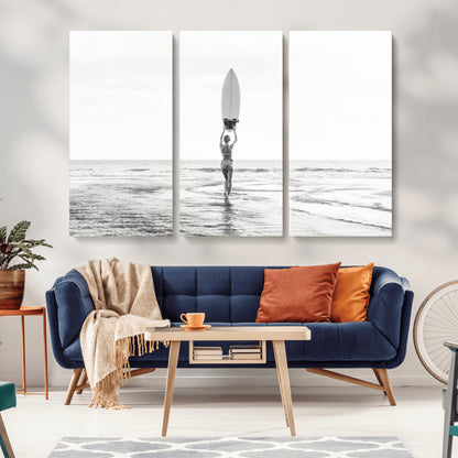 Black and white image of a female surfer holding a surfboard above her head while walking toward the ocean, creating a minimalist and bold coastal aesthetic.