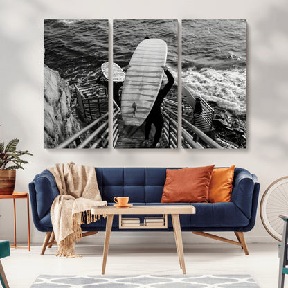Black and white photo of surfers carrying boards down a rugged cliffside stairway, capturing a bold and adventurous coastal scene.