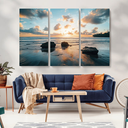 A serene beach at sunset with vibrant clouds, calm water, and rocks in the foreground reflecting the warm colors of the setting sun.