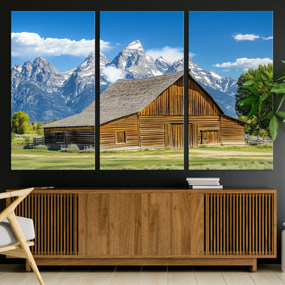 A rustic wooden barn sits in front of towering mountain peaks under a clear blue sky, perfect for farmhouse or cabin style interiors.