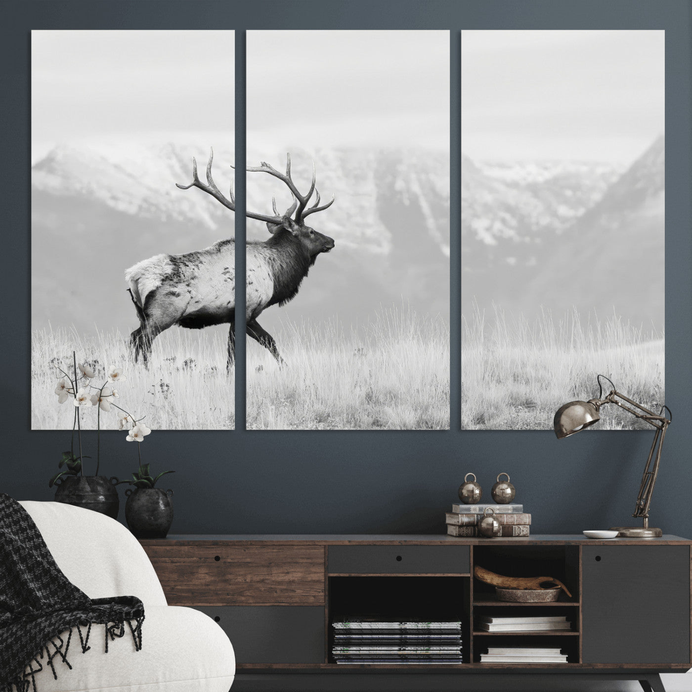 Black and white photograph of a majestic elk standing in a grassy field with snow-capped mountains in the background, creating a bold and rustic wilderness scene.