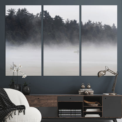 A lone surfer carrying a board through a misty shoreline, surrounded by dense trees and fog, creating a peaceful and mysterious coastal scene.