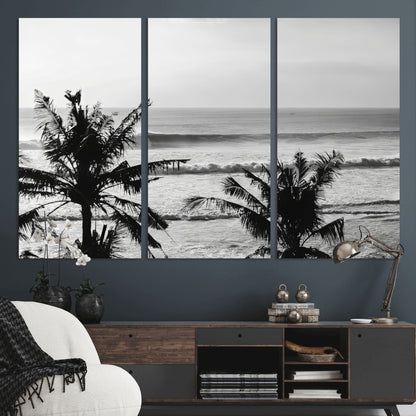 Black and white view of ocean waves crashing on a tropical coastline, framed by silhouetted palm trees under a vast, open sky.