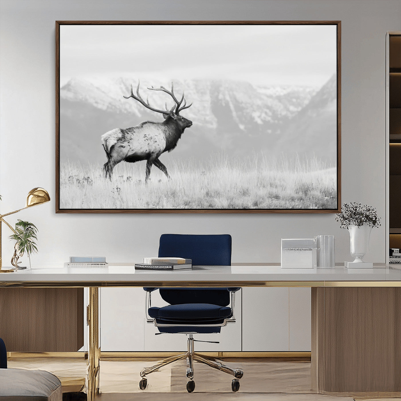Black and white photograph of a majestic elk standing in a grassy field with snow-capped mountains in the background, creating a bold and rustic wilderness scene.