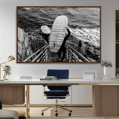 Black and white photo of surfers carrying boards down a rugged cliffside stairway, capturing a bold and adventurous coastal scene.