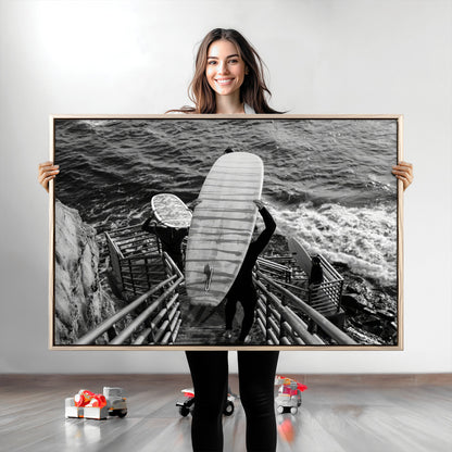 Black and white photo of surfers carrying boards down a rugged cliffside stairway, capturing a bold and adventurous coastal scene.