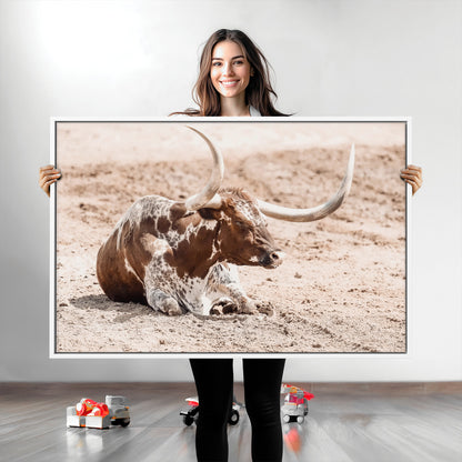 Texas Longhorn bull resting on dusty ground with warm tones, ideal for rustic western or farmhouse-themed wall decor.