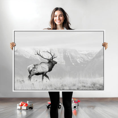 Black and white photograph of a majestic elk standing in a grassy field with snow-capped mountains in the background, creating a bold and rustic wilderness scene.