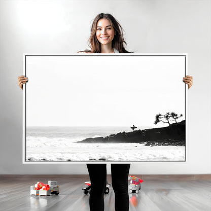 Black and white photo of a surfer standing on rocky cliffs, gazing at the waves, embodying the spirit of adventure and solitude.