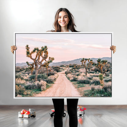 A dusty road winding through Joshua Tree National Park, framed by iconic twisted trees and warm desert tones under a soft pastel sky.