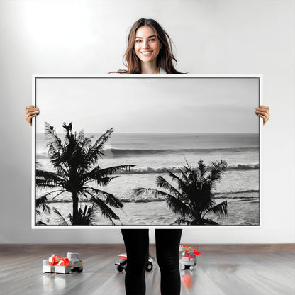 Black and white view of ocean waves crashing on a tropical coastline, framed by silhouetted palm trees under a vast, open sky.