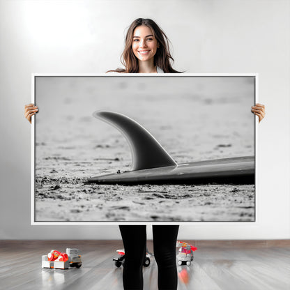 A black and white close-up of a surfboard fin resting on the sandy shore, highlighting minimalist beach aesthetics and classic surf culture.