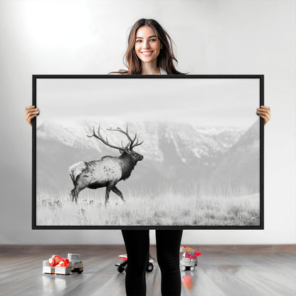 Black and white photograph of a majestic elk standing in a grassy field with snow-capped mountains in the background, creating a bold and rustic wilderness scene.