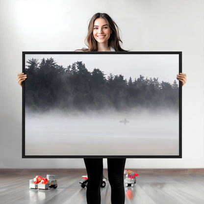 A lone surfer carrying a board through a misty shoreline, surrounded by dense trees and fog, creating a peaceful and mysterious coastal scene.