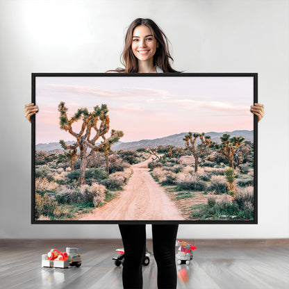 A dusty road winding through Joshua Tree National Park, framed by iconic twisted trees and warm desert tones under a soft pastel sky.