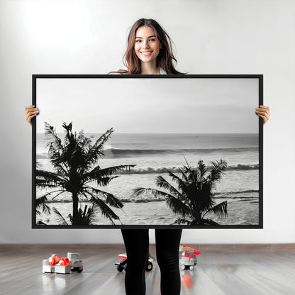 Black and white view of ocean waves crashing on a tropical coastline, framed by silhouetted palm trees under a vast, open sky.