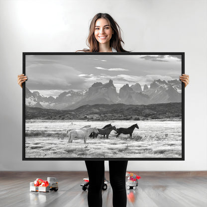 A black and white landscape of wild horses grazing in a vast grassland with towering mountains in the background, creating a moody and rustic western aesthetic.