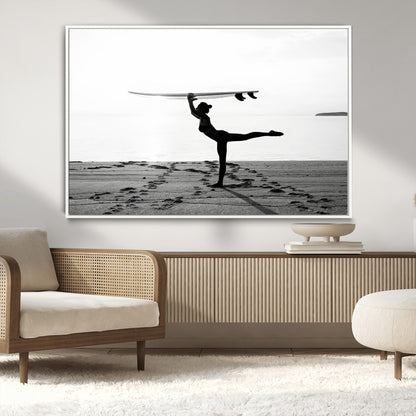 A woman balances a surfboard on her head while striking a yoga pose on the beach, silhouetted against a calm ocean horizon.