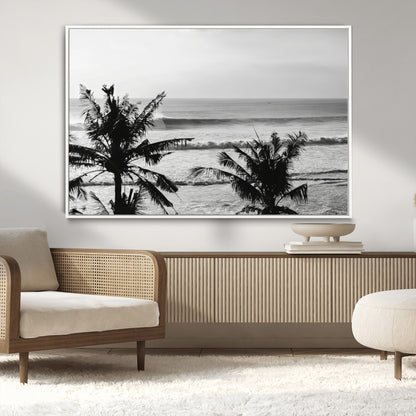 Black and white view of ocean waves crashing on a tropical coastline, framed by silhouetted palm trees under a vast, open sky.
