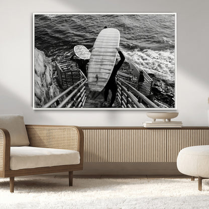 Black and white photo of surfers carrying boards down a rugged cliffside stairway, capturing a bold and adventurous coastal scene.