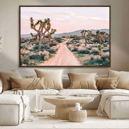 A dusty road winding through Joshua Tree National Park, framed by iconic twisted trees and warm desert tones under a soft pastel sky.