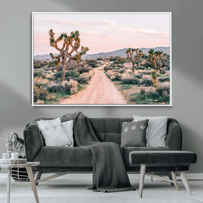 A dusty road winding through Joshua Tree National Park, framed by iconic twisted trees and warm desert tones under a soft pastel sky.