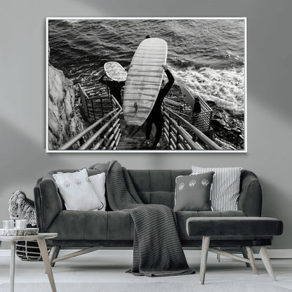 Black and white photo of surfers carrying boards down a rugged cliffside stairway, capturing a bold and adventurous coastal scene.