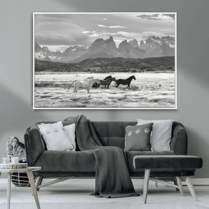 A black and white landscape of wild horses grazing in a vast grassland with towering mountains in the background, creating a moody and rustic western aesthetic.